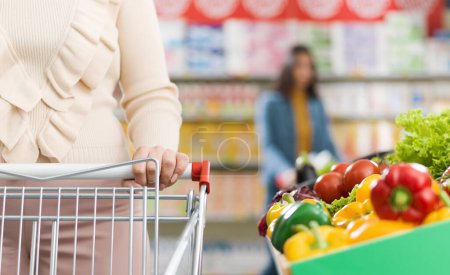 Téléchargez les photos : Légumes frais dans la section des produits au supermarché et femme poussant un panier - en image libre de droit