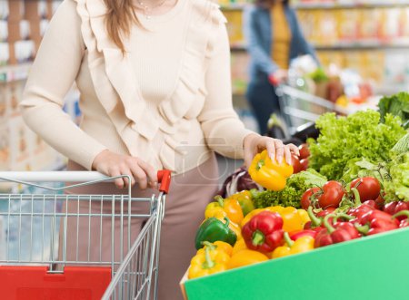 Téléchargez les photos : Femme poussant un chariot et achetant des légumes frais au supermarché, elle cueille un poivron - en image libre de droit