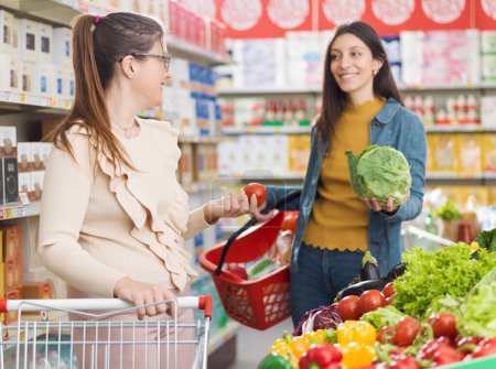 Foto de Mujeres felices que se reúnen en el supermercado, que están hablando y comprando verduras frescas y saludables juntos, compras de comestibles y el concepto de estilo de vida - Imagen libre de derechos