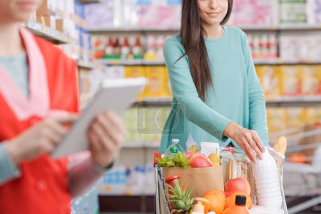 Téléchargez les photos : Femme poussant un panier complet au supermarché et commis de stock au travail, concept intérieur de supermarché - en image libre de droit