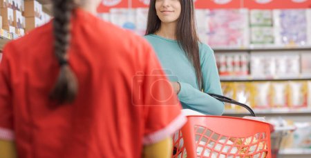 Smiling woman buying groceries at the supermarket, she is holding a shopping basket and talking with a friendly shop assistant, grocery shopping concept