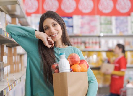 Foto de Retrato de una joven feliz sosteniendo una bolsa llena de comestibles en el supermercado, ella sonríe a la cámara - Imagen libre de derechos