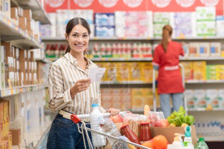 Porträt einer jungen lächelnden Frau beim Einkaufen im Supermarkt, sie hält eine Einkaufsliste in der Hand und schiebt einen vollen Einkaufswagen