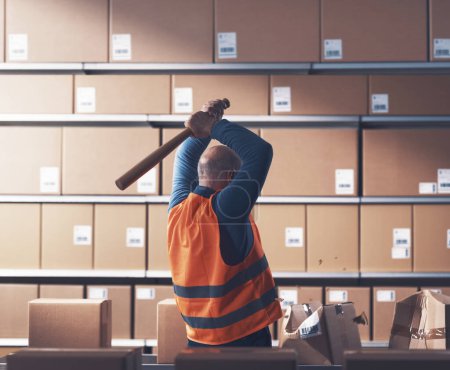 Crazy rebellious warehouse worker smashing cardboard boxes with a baseball bat