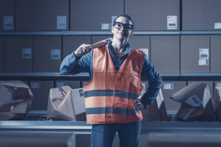 Photo for Confident smiling rebellious worker posing with a baseball bat at the warehouse, she is angry and she smashed many cardboard boxes - Royalty Free Image