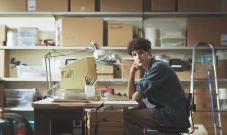 Téléchargez les photos : Adolescent intelligent travaillant avec un ordinateur vintage dans le garage, il regarde la caméra - en image libre de droit