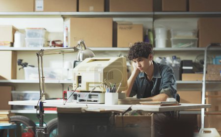 Téléchargez les photos : Ennui paresseux adolescent gars assis au bureau dans le garage et en utilisant un vieil ordinateur vintage - en image libre de droit
