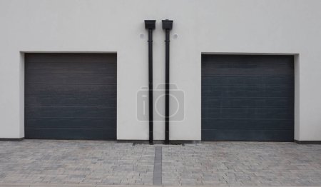 Dos puertas de garaje con persiana enrollable gris en fachada blanca. Liquidación en pavimento, camino de entrada.