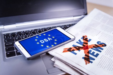 Smartphone con DSA y bandera de la UE en pantalla sobre un periódico con noticias falsas y un portátil. La Ley de Servicios Digitales de la Unión Europea o DSA regula la moderación proactiva, incluida la comprobación de los hechos