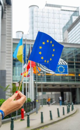 Plan rapproché d'une main tenant un petit drapeau de l'Union européenne devant le bâtiment du Parlement européen à Bruxelles, en Belgique. L'arrière-plan comporte plusieurs autres drapeaux et l'architecture moderne de