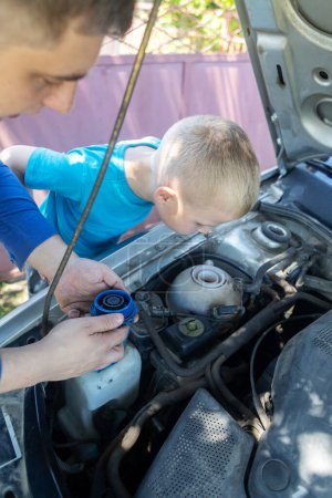 Photo for Dad shows his son where to fill in the coolant in the engine compartment. Topping up antifreeze in the car cooling system. Expansion tank near the engine. Red liquid - Royalty Free Image