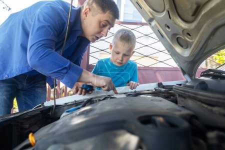 Photo for Dad shows his son where to fill in the coolant in the engine compartment. Topping up antifreeze in the car cooling system. Expansion tank near the engine. Red liquid - Royalty Free Image
