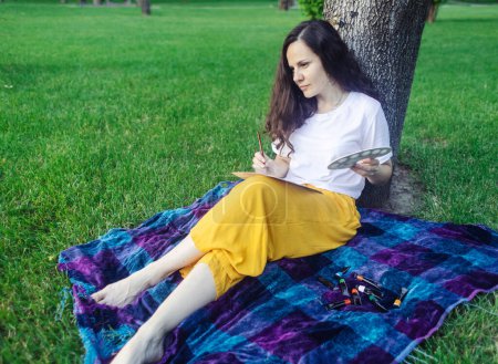 Photo for Art therapy. Woman draws in the park. Girl sits with her back against a tree and looks into distance. Restoration of nervous system with help of drawing. Concept of relaxation and meditative state - Royalty Free Image