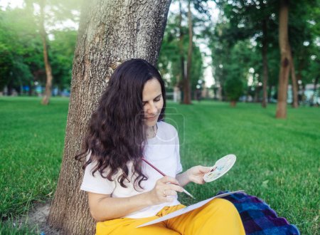 Photo for Art therapy. Woman draws in the park. Girl sits with her back against a tree and looks into distance. Restoration of nervous system with help of drawing. Concept of relaxation and meditative state - Royalty Free Image