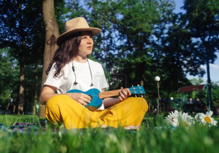 Foto de Una chica en el parque sentada en la hierba toca el ukelele. Aprender a tocar un instrumento de cuatro cuerdas. Relajación en un parque público. Musicoterapia, psicología, meditación, relajación, relajación - Imagen libre de derechos