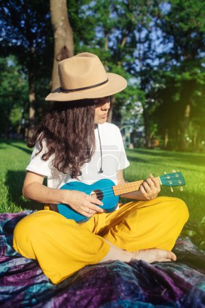 Foto de Una chica en el parque sentada en la hierba toca el ukelele. Aprender a tocar un instrumento de cuatro cuerdas. Relajación en un parque público. Musicoterapia, psicología, meditación, relajación, relajación - Imagen libre de derechos