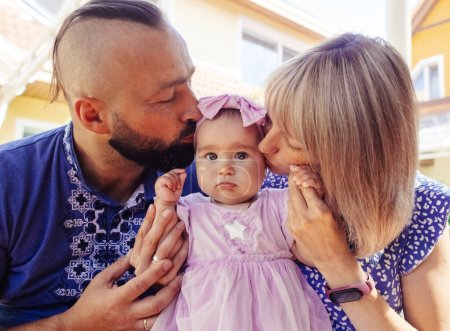 Eltern in ukrainischen Stickereien küssen ihr Kind. Ein kleines Mädchen mit dunklen Augen und ihre liebevolle Mutter und ihr Vater. Das Konzept einer glücklichen Kindheit und die Manifestation von Fürsorge.