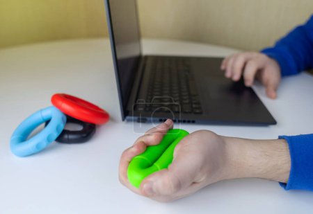 Hand gripper. Man squeezes rubber expander while working on his laptop. Concept of combining useful activity with necessary work. Training hand and arm muscles. Multi-colored mini training apparatus