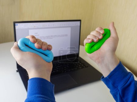 Pinza de mano. El hombre aprieta el expansor de goma mientras trabaja en su computadora portátil. Concepto de combinar la actividad útil con el trabajo necesario. Entrenamiento de músculos de mano y brazo. Mini aparato de entrenamiento multicolor