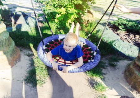 Happy fair-haired boy rides on nest swing or spider web. Cute portrait of a child on the playground in the park. A game in the summer outdoors
