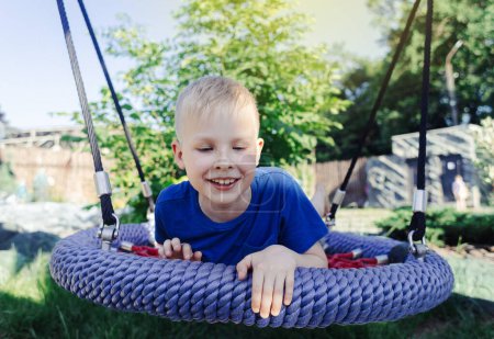 Happy fair-haired boy rides on nest swing or spider web. Cute portrait of a child on the playground in the park. A game in the summer outdoors