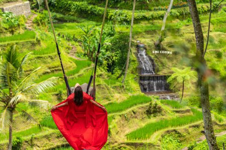 Photo for Tegalalang, Ubud, Indonasia - February 14.2023: Backview of a woman in long red dress on a swing at the natural terrace swing fairground rice field - Royalty Free Image