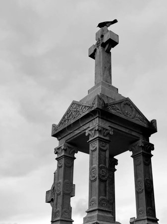Photo for Black and white picture of a graveyard monument. A cross watches over, as a black bird, a raven observes, casting an eerie yet mesmerizing scene. - Royalty Free Image