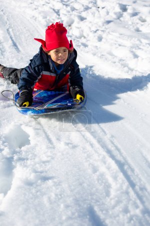 Téléchargez les photos : Joli garçon afro-américain qui descend la colline par une journée enneigée. Amusement hivernal dans la neige. Enfant divers sur un traîneau. Beaucoup d'espace de copie - en image libre de droit