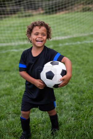 Photo for Young African American soccer player standing in front of a soccer goal smiling before a soccer game on a large grass field holding a ball - Royalty Free Image