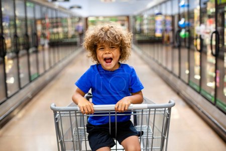 Téléchargez les photos : Petit garçon drôle riant assis dans un chariot lors d'un voyage en famille à l'épicerie. Il est excité d'acheter quelque chose de nouveau - en image libre de droit
