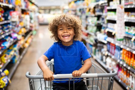 Foto de Divertido niño riéndose mientras está sentado en un carrito de compras durante un viaje familiar a la tienda de comestibles. Él está emocionado de comprar algo nuevo - Imagen libre de derechos