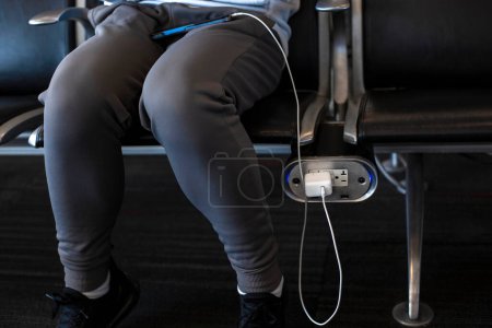 Téléchargez les photos : Image recadrée d'une femme assise dans un terminal de l'aéroport et chargeant un téléphone portable - en image libre de droit