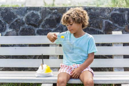 Foto de Lindo niño diverso comiendo un cono de nieve o afeitarse hielo al aire libre en un banco público de la calle. Disfrutando de un refresco fresco en un caluroso día de verano - Imagen libre de derechos