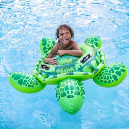 Téléchargez les photos : Mignon jeune garçon afro-américain souriant jouant joyeusement dans une piscine avec un jouet gonflable de tortue. Vue du dessus d'un garçon flottant et jouant dans l'eau - en image libre de droit