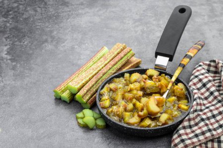 Homemade  rhubarb chutney in small skillet with raw stalk rhubarb on gray table 