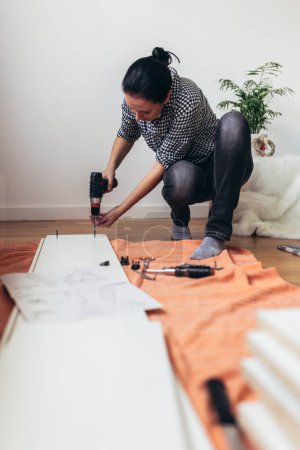 Téléchargez les photos : Femme en vêtements décontractés assis sur le sol de son appartement et assembler des meubles - en image libre de droit
