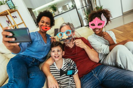 Photo for Happy family with party props taking selfie by smartphone sitting on sofa at home - Royalty Free Image