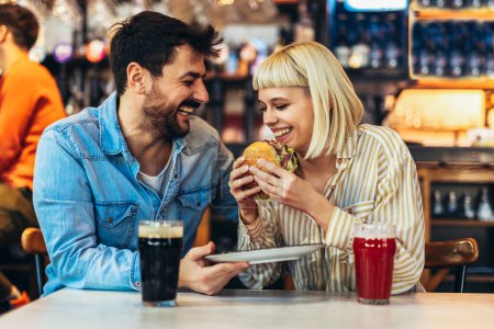 Photo for Young couple in love having fun spending leisure time together at restaurant, eating burgers and drinking beer - Royalty Free Image