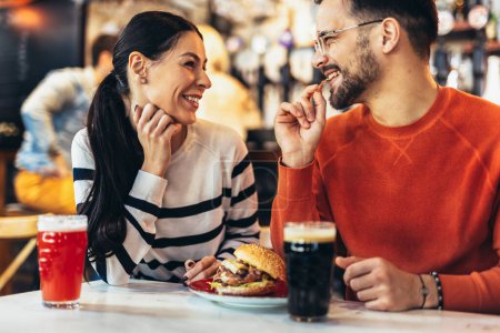 Photo for Young couple in love having fun spending leisure time together at restaurant, eating burgers and drinking beer - Royalty Free Image
