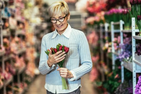 Téléchargez les photos : Vieille dame souriante, tenant un bouquet de fleurs à la main dans la boutique de fleurs. - en image libre de droit