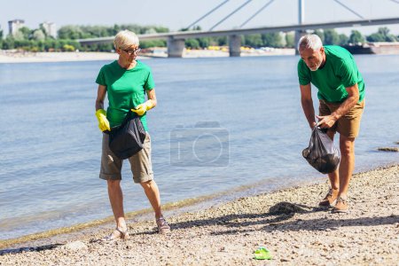 Photo for Mature volunteers gathering garbage on river bank. Ecology concept - Royalty Free Image