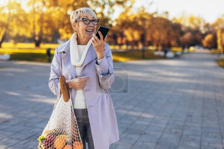 Foto de Feliz sonriente mujer mayor está sosteniendo una bolsa con frutas y verduras al aire libre utilizando un teléfono - Imagen libre de derechos