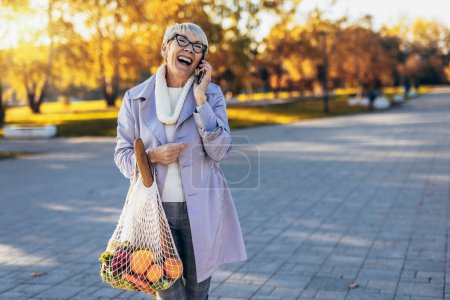 Foto de Feliz sonriente mujer mayor está sosteniendo una bolsa con frutas y verduras al aire libre utilizando un teléfono - Imagen libre de derechos