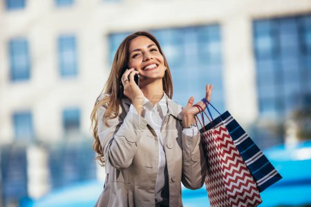 Foto de Mujer joven atractiva usando su teléfono celular afuera mientras hace compras en la ciudad - Imagen libre de derechos