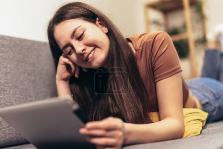 Foto de Young teen girl holding digital tablet computer looking at screen reading e book, watching movie video,make video call, surfing internet relaxing lying on sofa at home in living room. - Imagen libre de derechos
