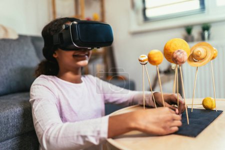 Foto de Happy african american school girl making a solar system for a school science project at home using VR glasses to immerse herself in metaverse - Imagen libre de derechos