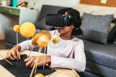 Photo for Happy african american school girl making a solar system for a school science project at home using VR glasses to immerse herself in metaverse - Royalty Free Image