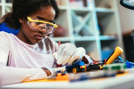 Foto de Smart Young African American Schoolgirl is Studying Electronics and Soldering Wires and Circuit Boards in Her Science Hobby Robotics Project. Girl is Working on a Robot in Her Room. Education Concept. - Imagen libre de derechos