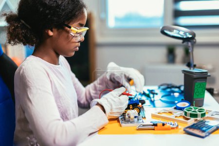 Téléchargez les photos : Smart Young African American Schoolgirl is Studying Electronics and Soldering Wires and Circuit Boards in Her Science Hobby Robotics Project. Girl is Working on a Robot in Her Room. Education Concept. - en image libre de droit