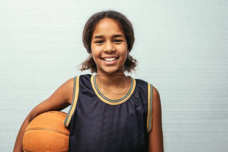 Foto de Female young basketball player standing against wall. Confident teenage girl is holding ball. She is wearing black jersey. - Imagen libre de derechos